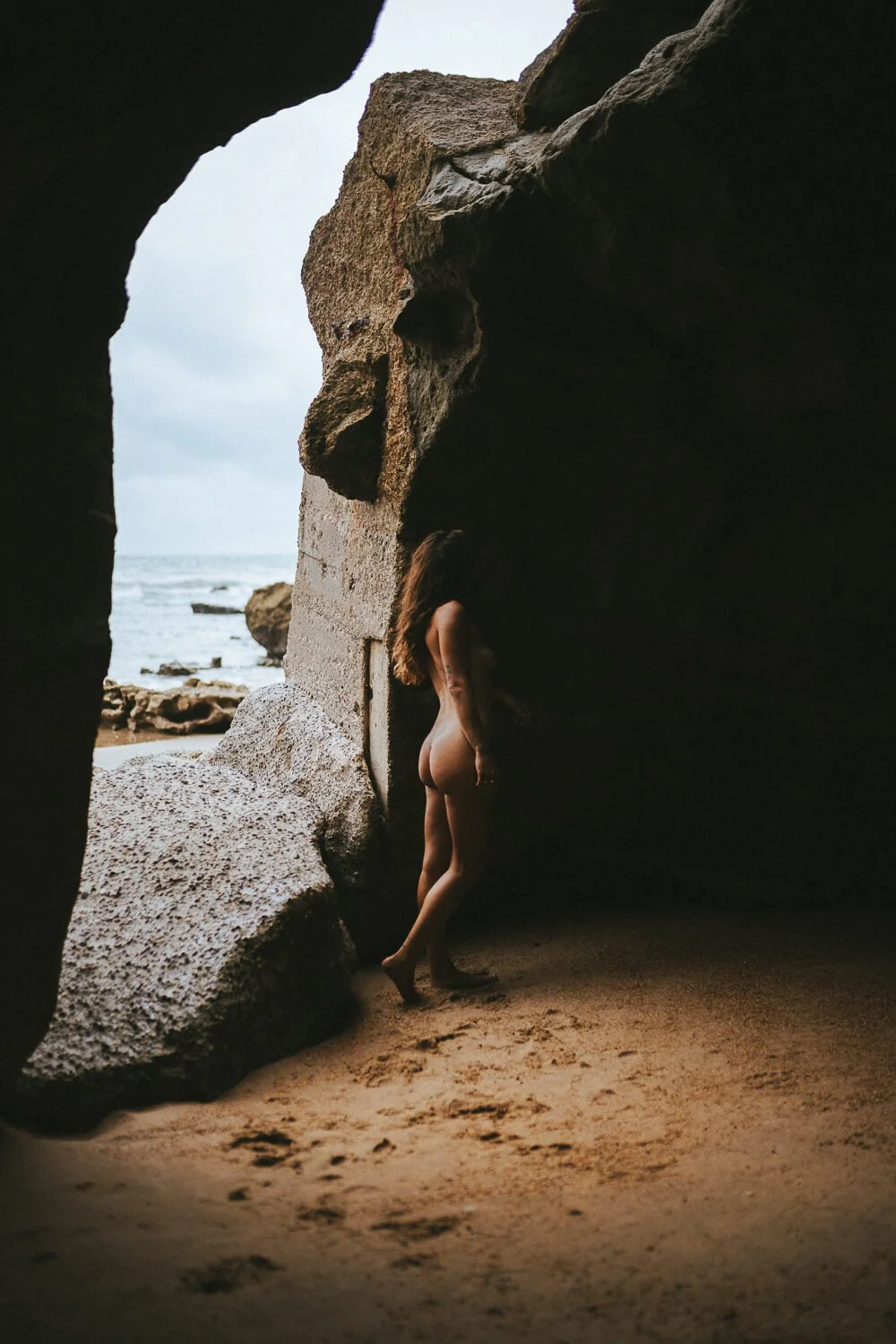 Nude woman standing inside a rocky cave at the beach, facing away from the camera, with the ocean visible in the background.