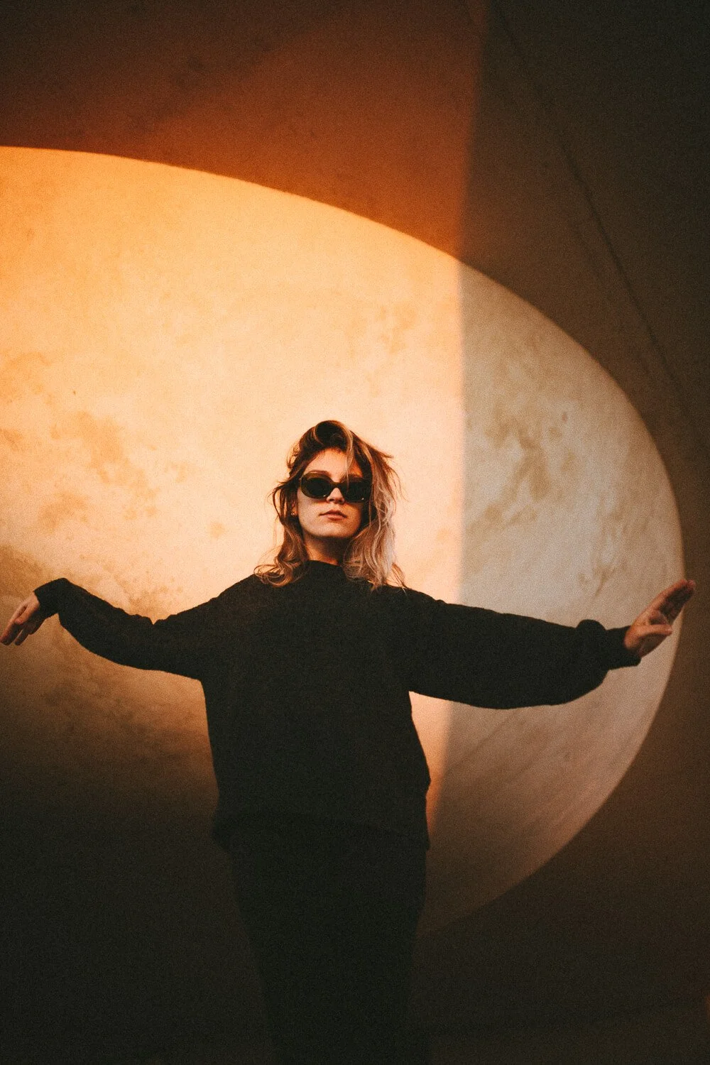 A woman with wavy hair wearing sunglasses and a black sweater standing with her arms outstretched in front of a large, glowing spherical art installation.