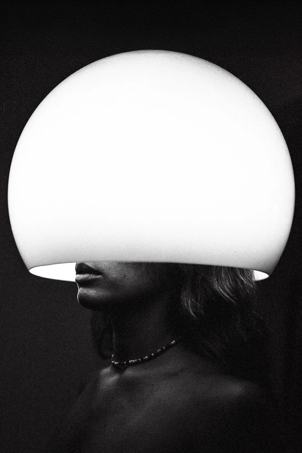Black and white photo of a woman with a large illuminated lampshade covering her eyes and forehead, showing only her nose, lips, and part of her neck and shoulder, with a necklace.