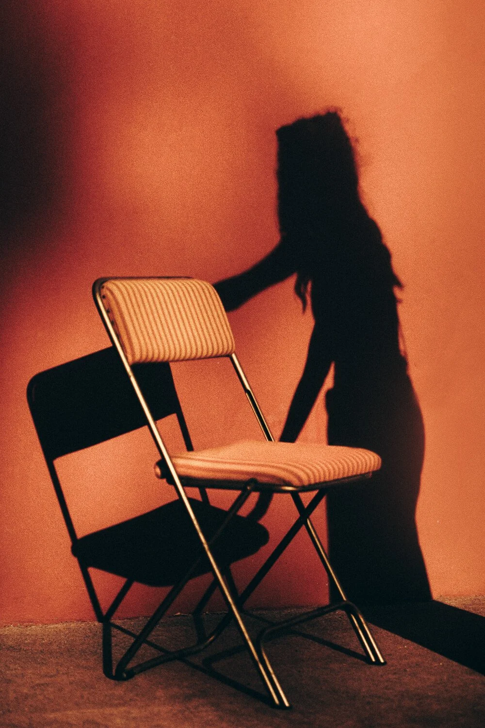 Shadow of a woman pushing a striped folding chair against a reddish wall.