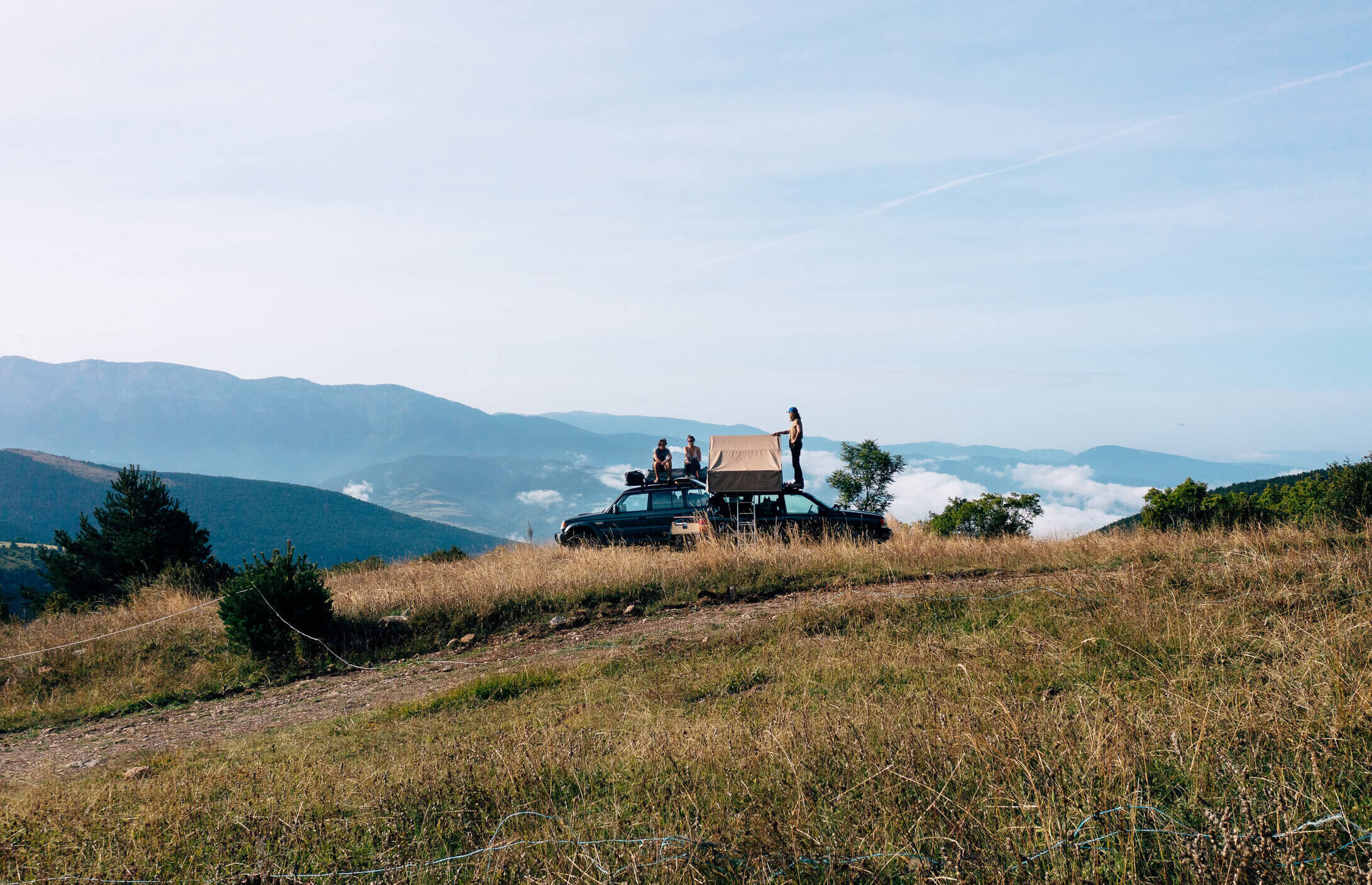 Quatre personnes sur une voiture avec une tente de toit, stationnée sur une colline avec vue panoramique sur les montagnes et un ciel clair.