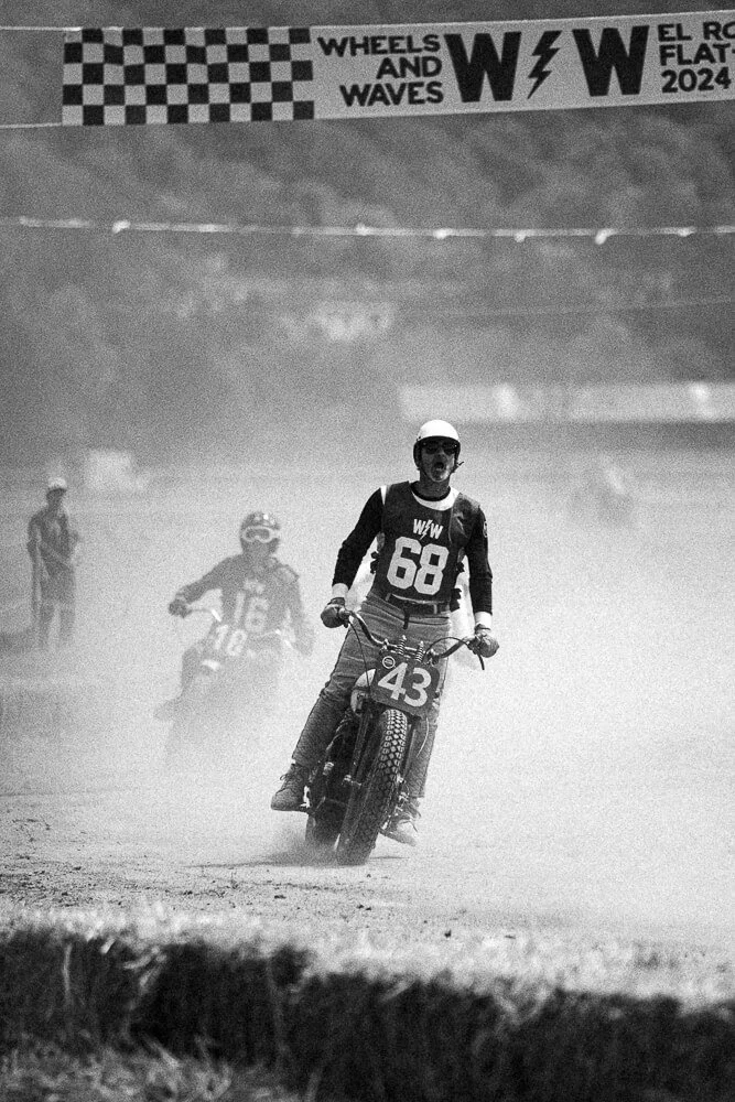 Two motocross racers riding on a dusty track, one in front wearing jersey number 43 and the other behind wearing jersey number 16, with a banner overhead reading 'WHEELS AND WAVES WW EL ROLLO FLOTT 2024'.