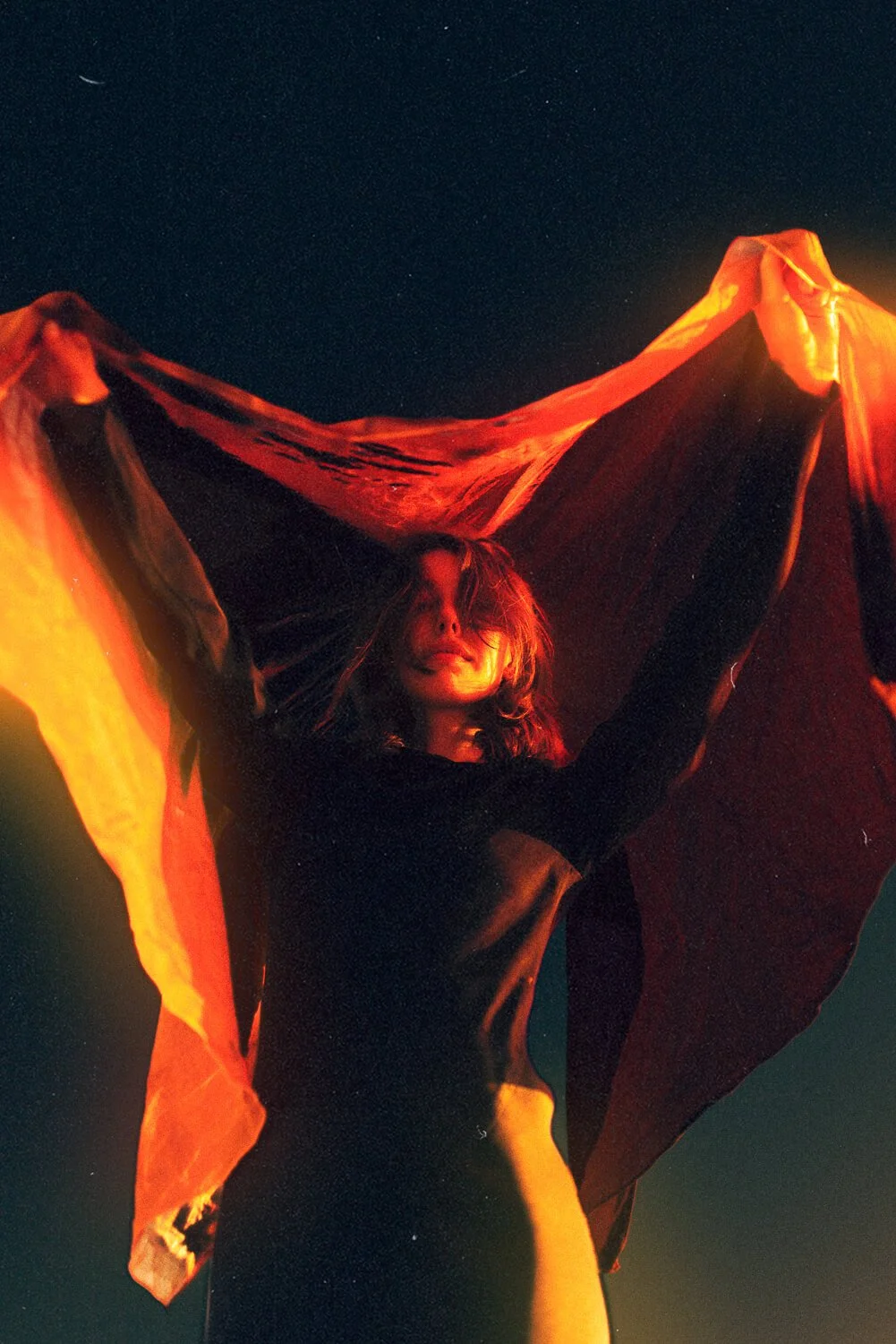 Woman standing outdoors at night holding a large, flowing red and orange fabric above her head, with a starry sky in the background.