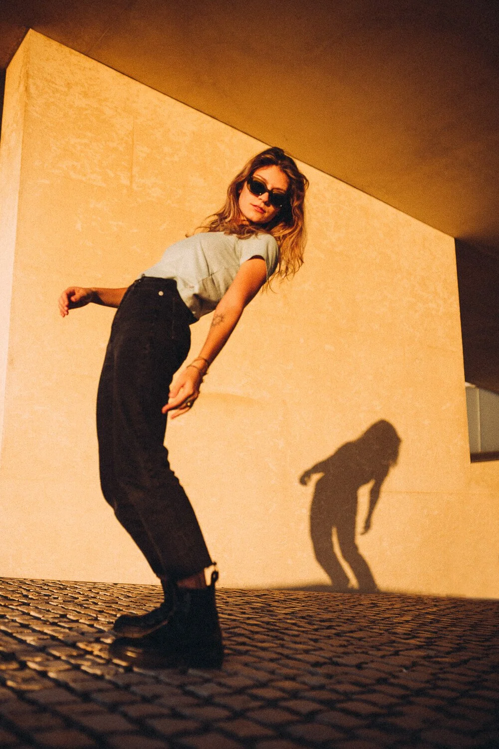 A woman with wavy hair wearing sunglasses, a light-colored t-shirt, ripped black jeans, and black boots, is posing outdoors during sunset, casting a long shadow on a textured yellow wall.