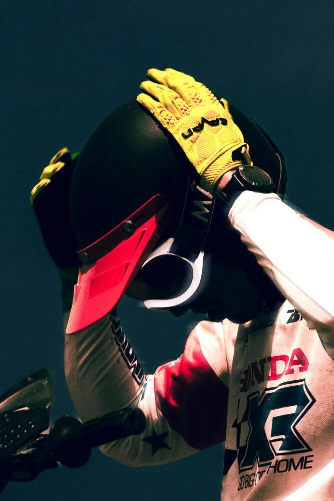 A person wearing a white racing suit with red and black accents, yellow gloves, and a black helmet with a red visor, holding the helmet with both hands against a dark background.