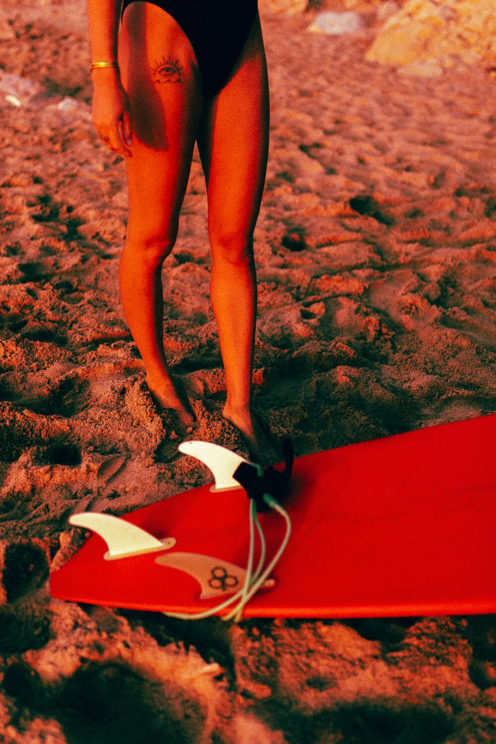 A person standing on sandy ground in a swimming pool or beach area, wearing black swimwear and a gold bracelet, with a tattoo of an eye on their upper thigh. Part of a red surfboard with white fins and a leash is visible in the foreground.