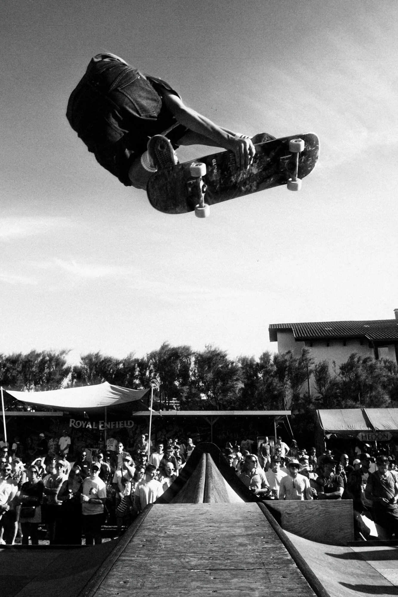 A skateboarder in mid-air performing a jump at an outdoor event with a crowd and tents below.