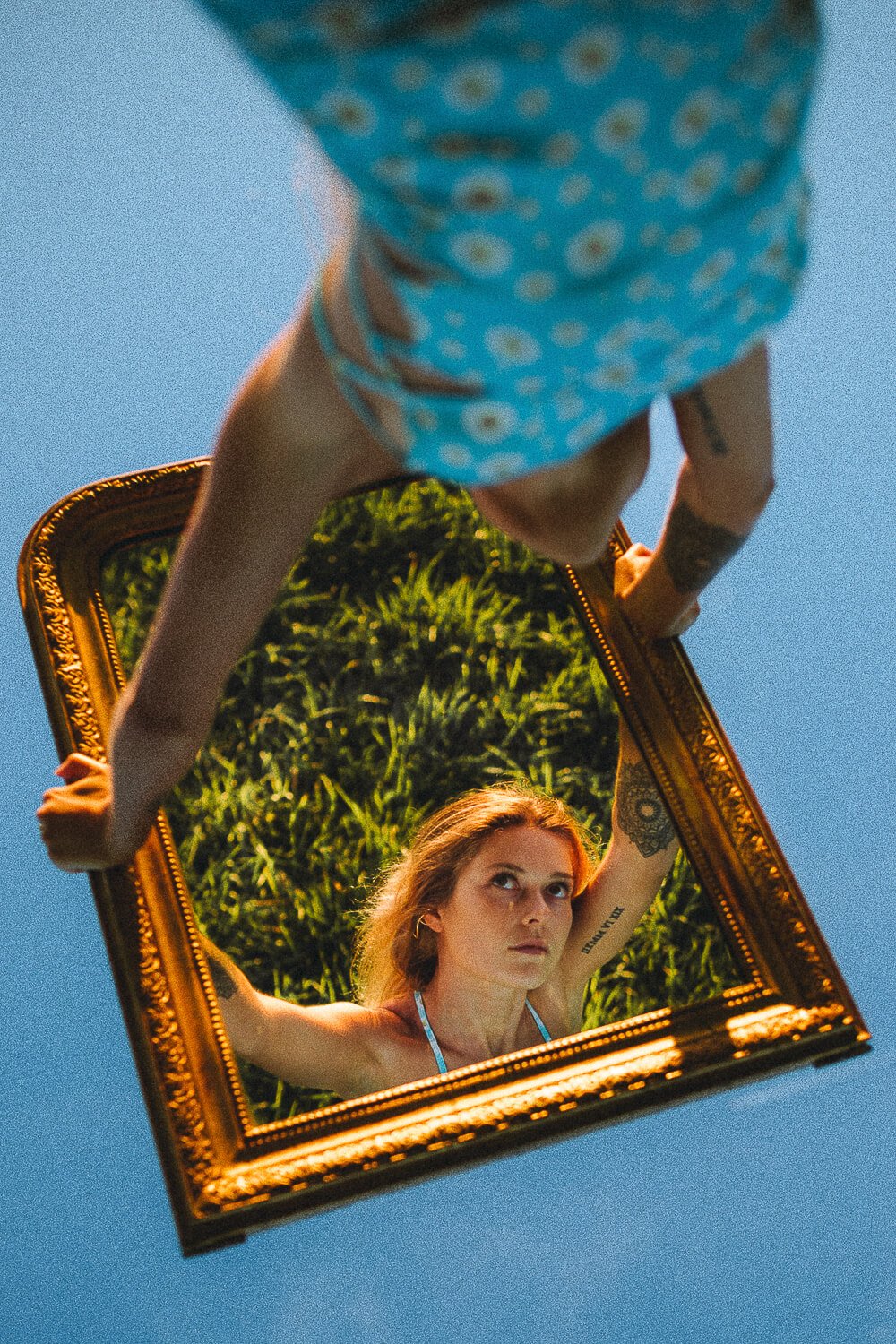 A woman with red hair and tattoos looks into a mirror reflecting her face and upper body, held outdoors against a blue sky and greenery.
