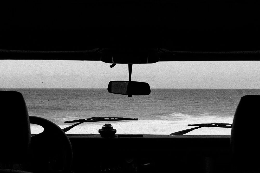 View from inside a vehicle, looking through the windshield at the ocean with waves, under a cloudy sky.