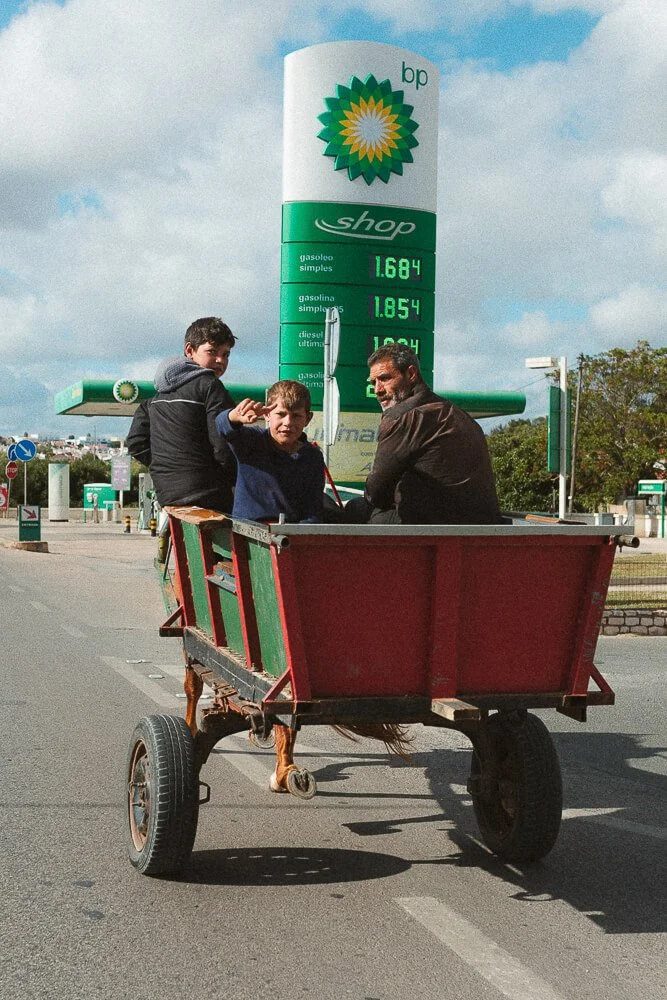 Three people riding in a red and green cart on an empty street outside a BP gas station with fuel prices displayed, under a partly cloudy sky.