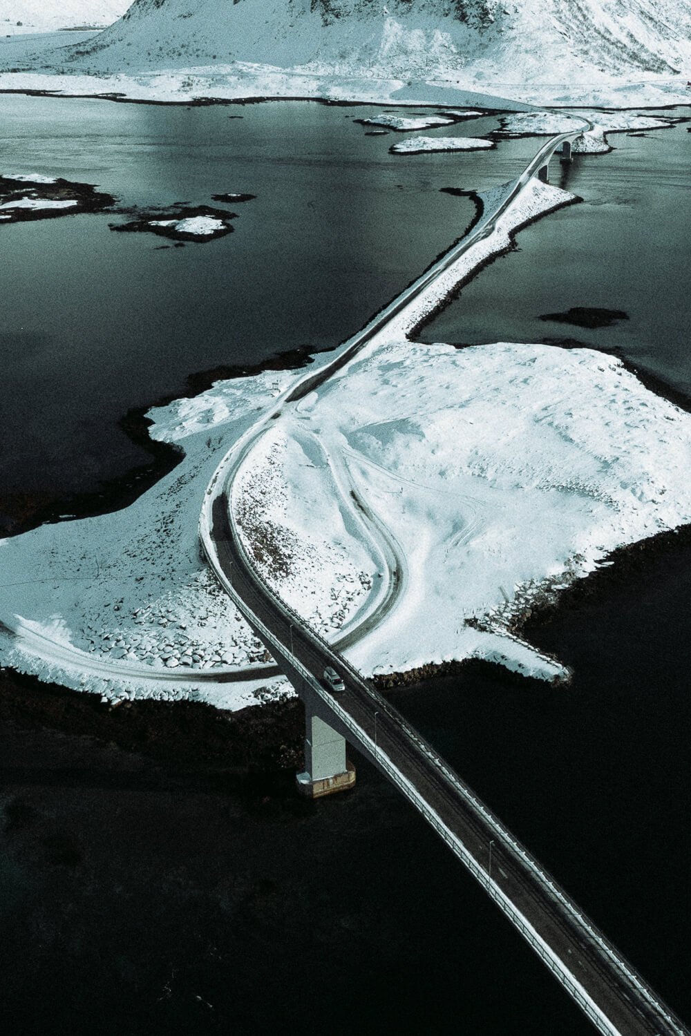Un pont long et sinueux traversant une région enneigée et un lac, avec des îles et des glaciers au fond.