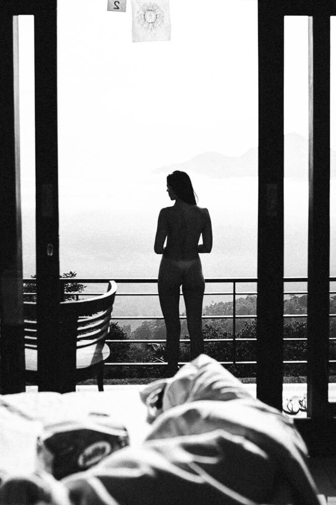 A woman standing on a balcony, looking out at a misty landscape with mountains, seen from inside a room with a bed and a chair.