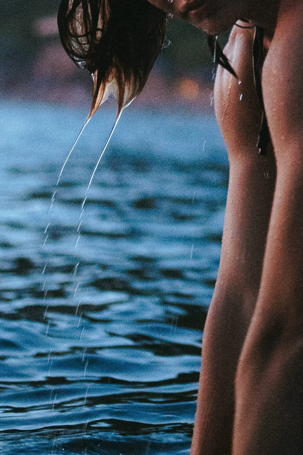A person with wet hair leans over water, with water dripping from hair and body, in a close-up shot.