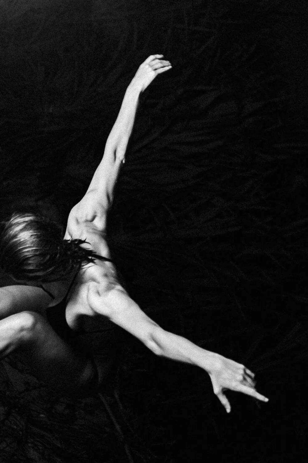 A black and white photograph of a woman in a dynamic pose, with one arm extended and her head down, against a dark background with textured patterns.