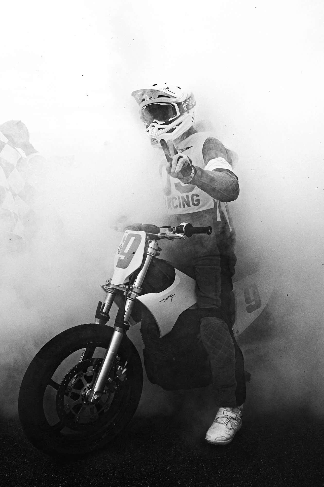 A person in motocross gear standing next to a motocross bike, giving a thumbs-up gesture while surrounded by a cloud of smoke.