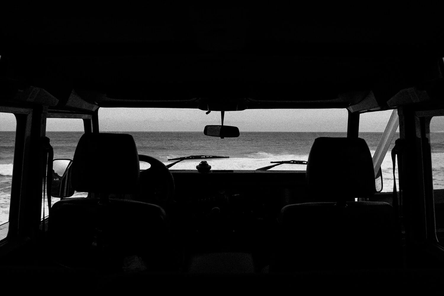 View from inside a vehicle facing the ocean, with the dashboard and seats visible in silhouette.