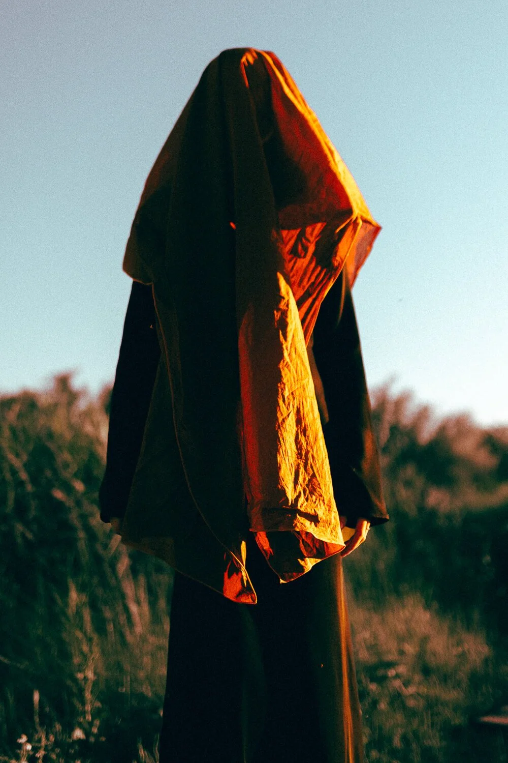 Person standing outdoors with a jacket draped over their head during sunset, with trees in the background.
