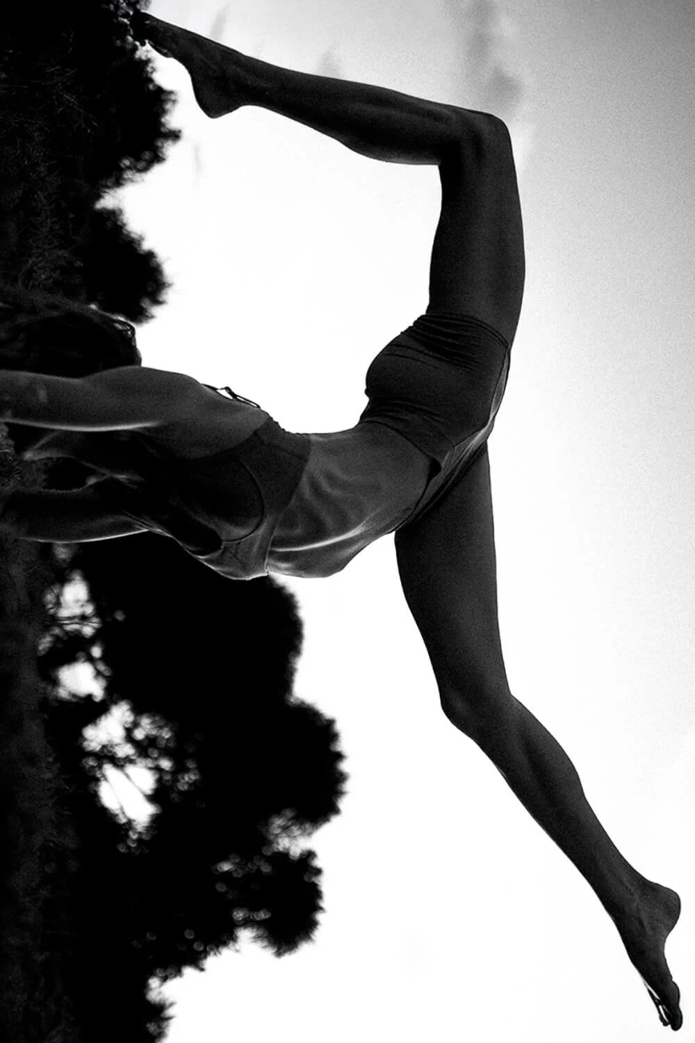 A person performing a yoga pose outdoors, silhouetted against the sky in black and white. The person is balancing on one leg with the other leg extended backward and arms reaching forward.