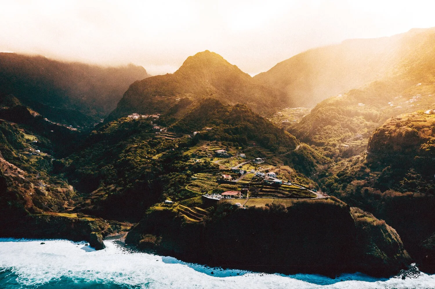 Scenic view of lush green mountains with terraced fields and scattered houses near the coast, with ocean waves crashing at the shoreline, under a partly cloudy sky with sunlight illuminating the landscape.