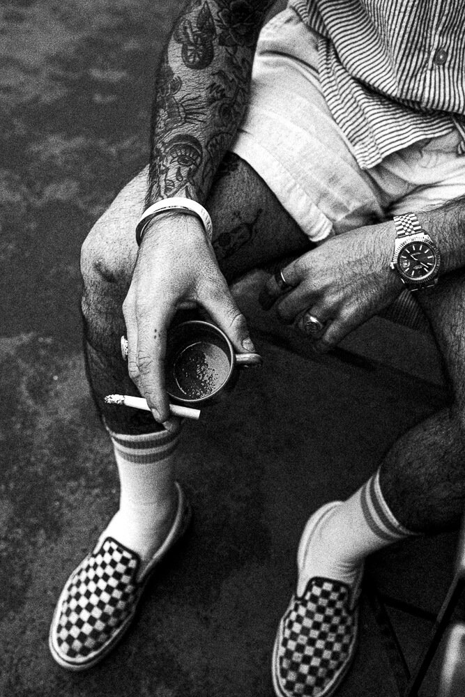 A person sitting down, holding a cup of coffee or tea with a cigarette in hand. They are wearing checkered slip-on shoes, striped socks, shorts, and a striped shirt. The person has tattoos on their arm and is wearing a watch and rings.