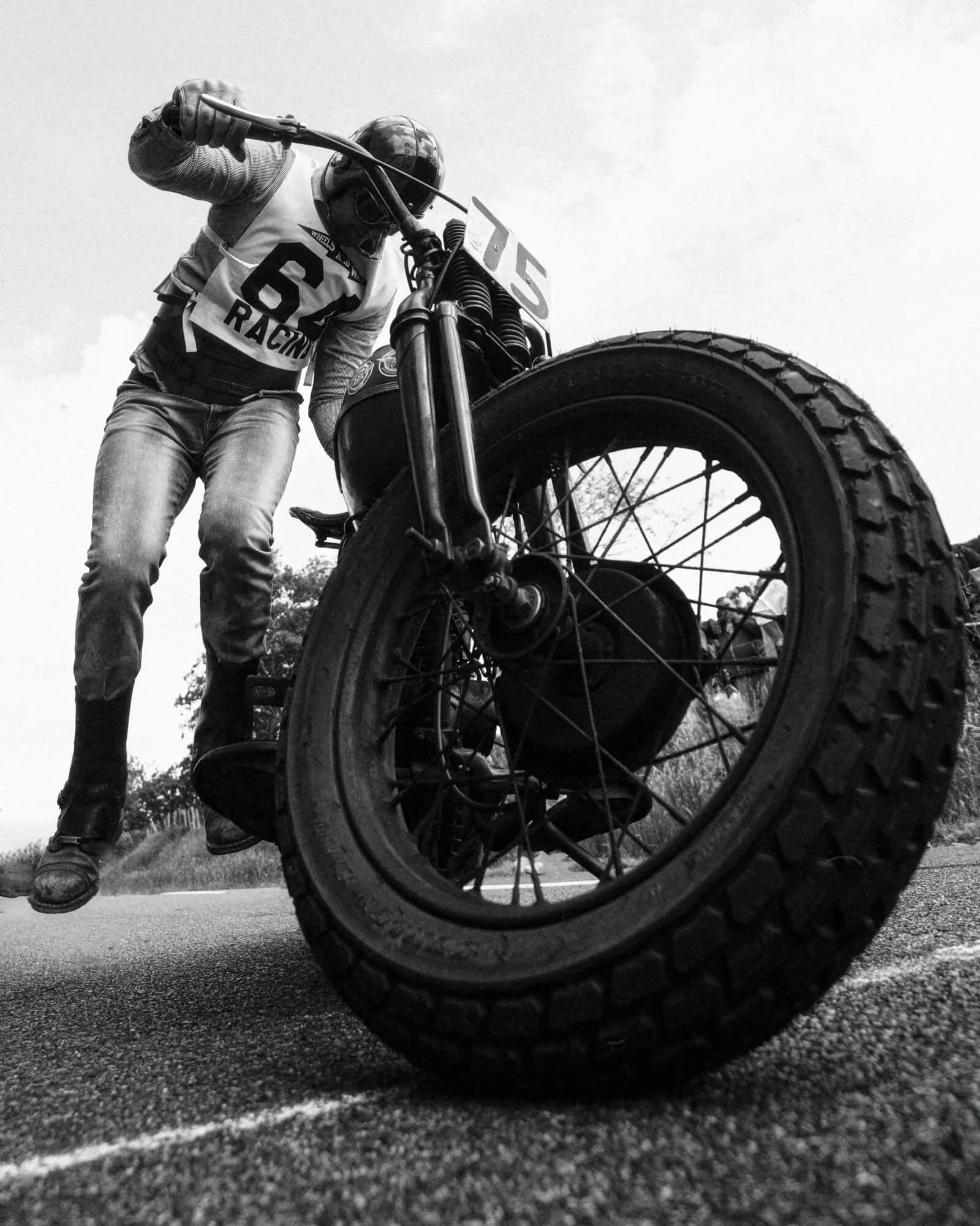 Black and white photo of a person inspecting a motorcycle tire from underneath, wearing a helmet and riding gear, with the motorcycle's front wheel in the foreground.