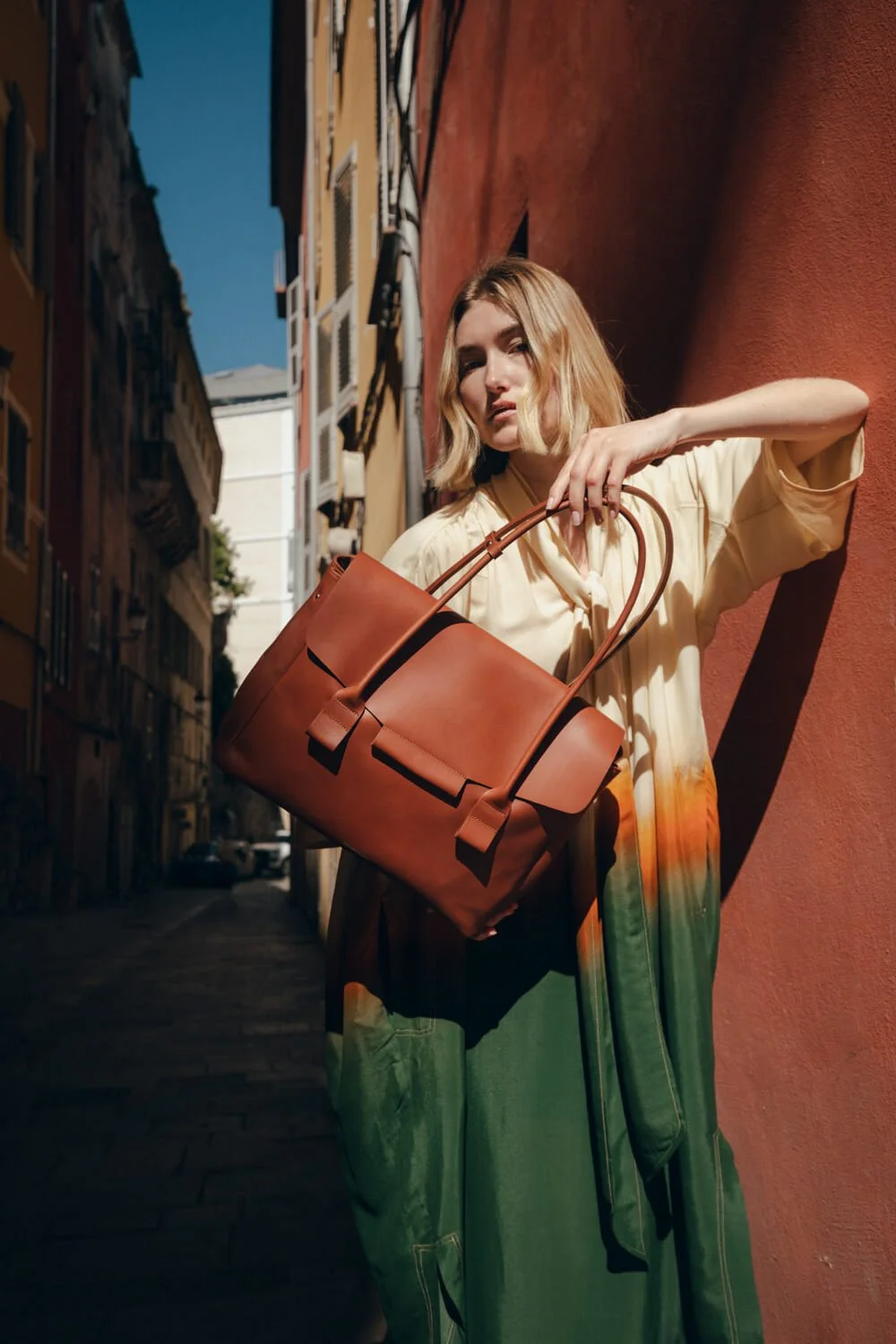 A woman with shoulder-length blonde hair standing against a red wall in a narrow street, holding a large brown leather bag.