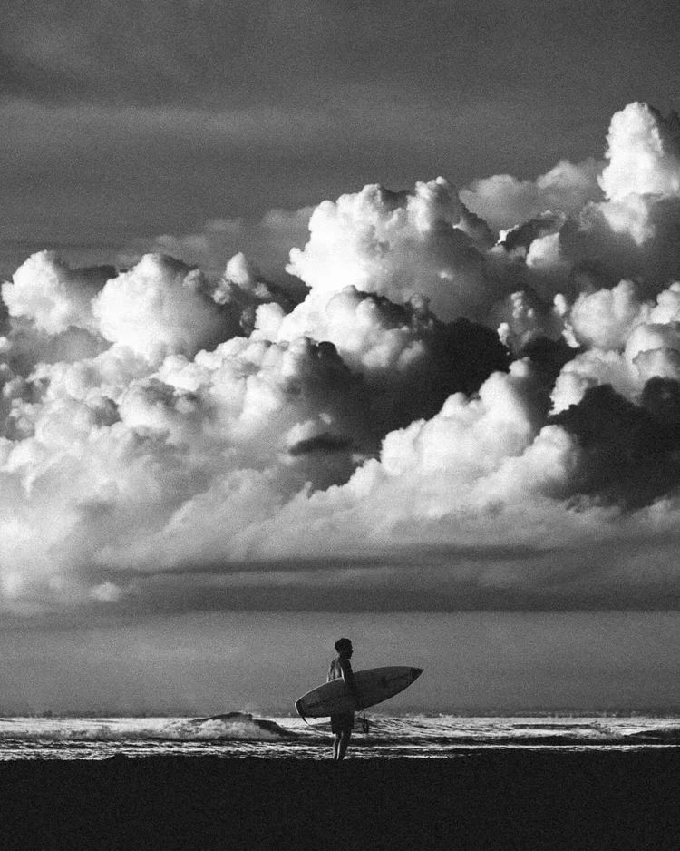 Une personne debout sur la plage avec une planche de surf, regardant vers la mer sous un ciel orageux avec de grands nuages.