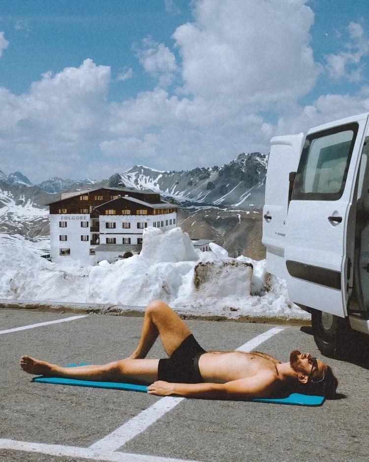 Hommes bronzant en maillot de bain sur un tapis de yoga dans un parking de montagne avec neige, à côté d'un van blanc, en arrière-plan des montagnes et un bâtiment hôtelier.