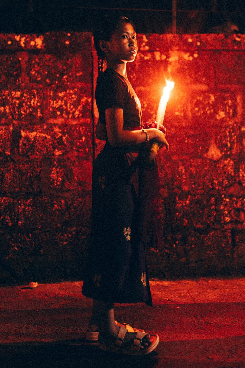 Jeune fille debout en noir, tenant une torche allumée, devant un mur en pierre, dans l'obscurité, ambiance nocturne chaleureuse.