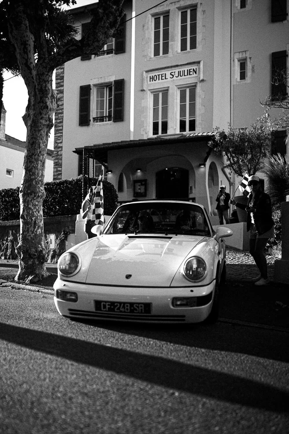 A vintage white Porsche parked in front of a hotel with the sign "Hotel St. Julien." There is a woman with a hat holding a checkered flag, and other people in the background near the hotel entrance. The scene is outdoors on a street with trees and bu