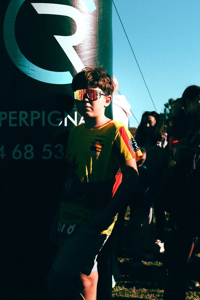 A young boy wearing sunglasses, a yellow sports jersey, and black shorts standing among a crowd outdoors, with a large black and green sign in the background.