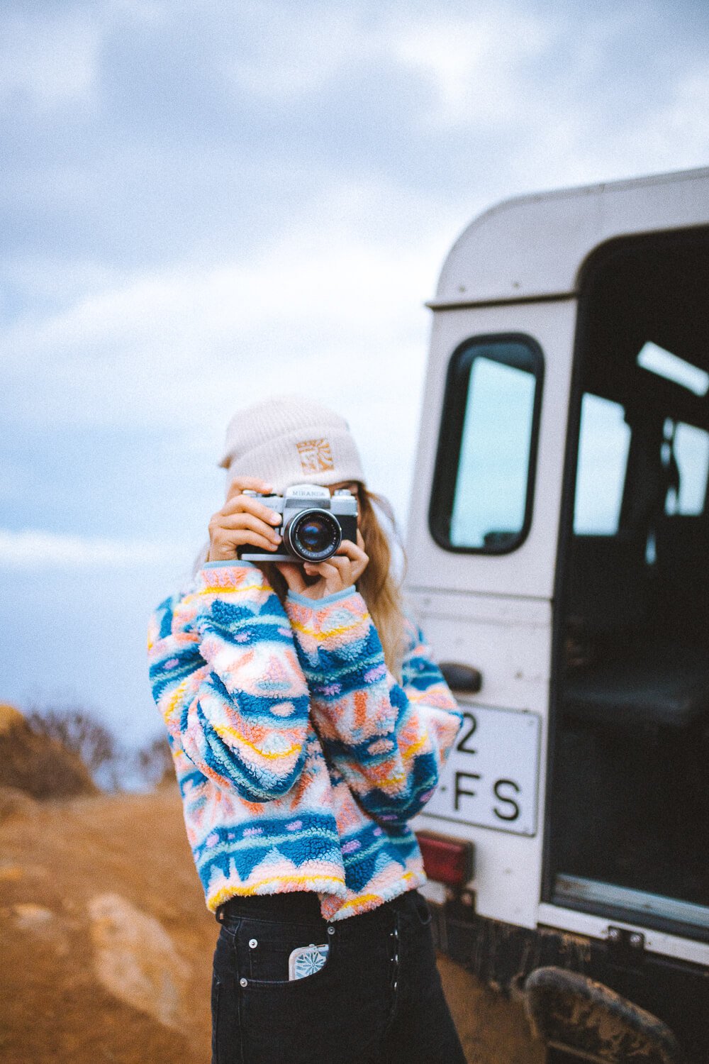 Person wearing a beige beanie, colorful fleece jacket, and black pants holds a camera up to take a picture outdoors near a white off-road vehicle with a license plate. The background shows a cloudy sky and dirt terrain.