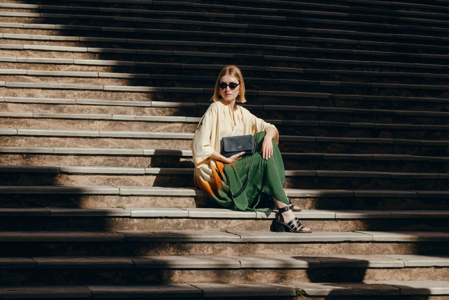 A woman with blonde hair sitting on outdoor steps, wearing sunglasses, a cream-colored blouse, green and orange patterned pants, and black heels, holding a black handbag.