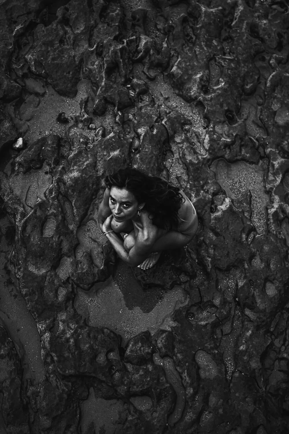 A woman with long hair crouching on rocky terrain, looking up at the camera in black and white.