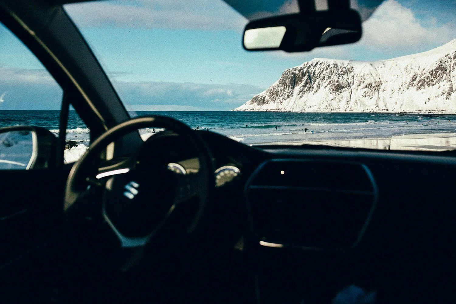 Vue depuis l'intérieur d'une voiture vers une plage avec des vagues et des montagnes enneigées au loin, en Norvège.