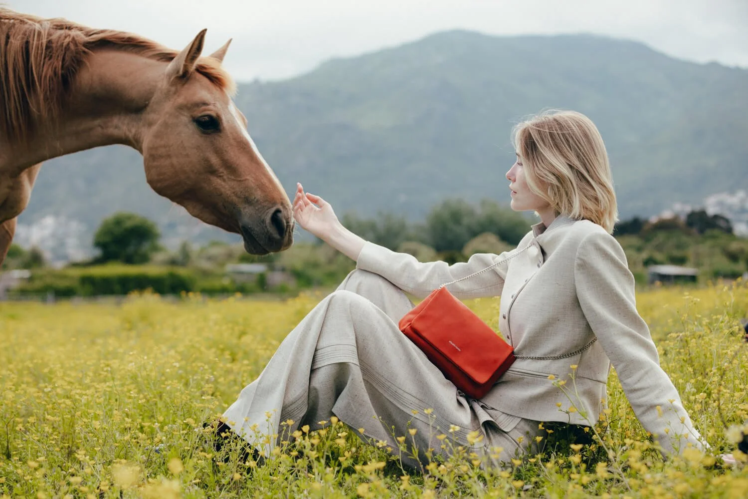 A woman with wavy blonde hair in a light-colored suit sitting on a field of yellow flowers, reaching out to a brown horse.