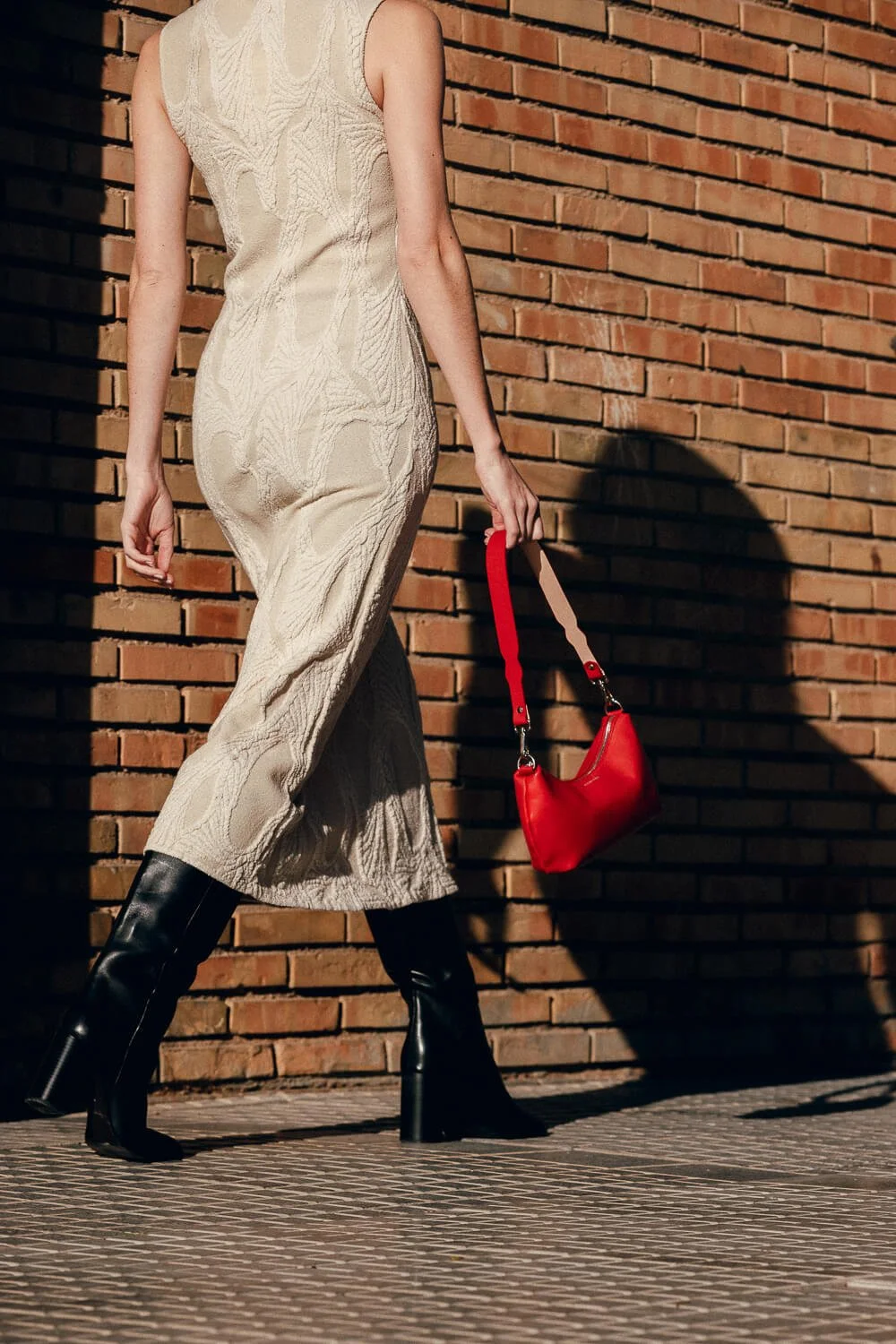 A woman walking against a brick wall, wearing a cream-colored textured dress, black knee-high boots, and carrying a red handbag.