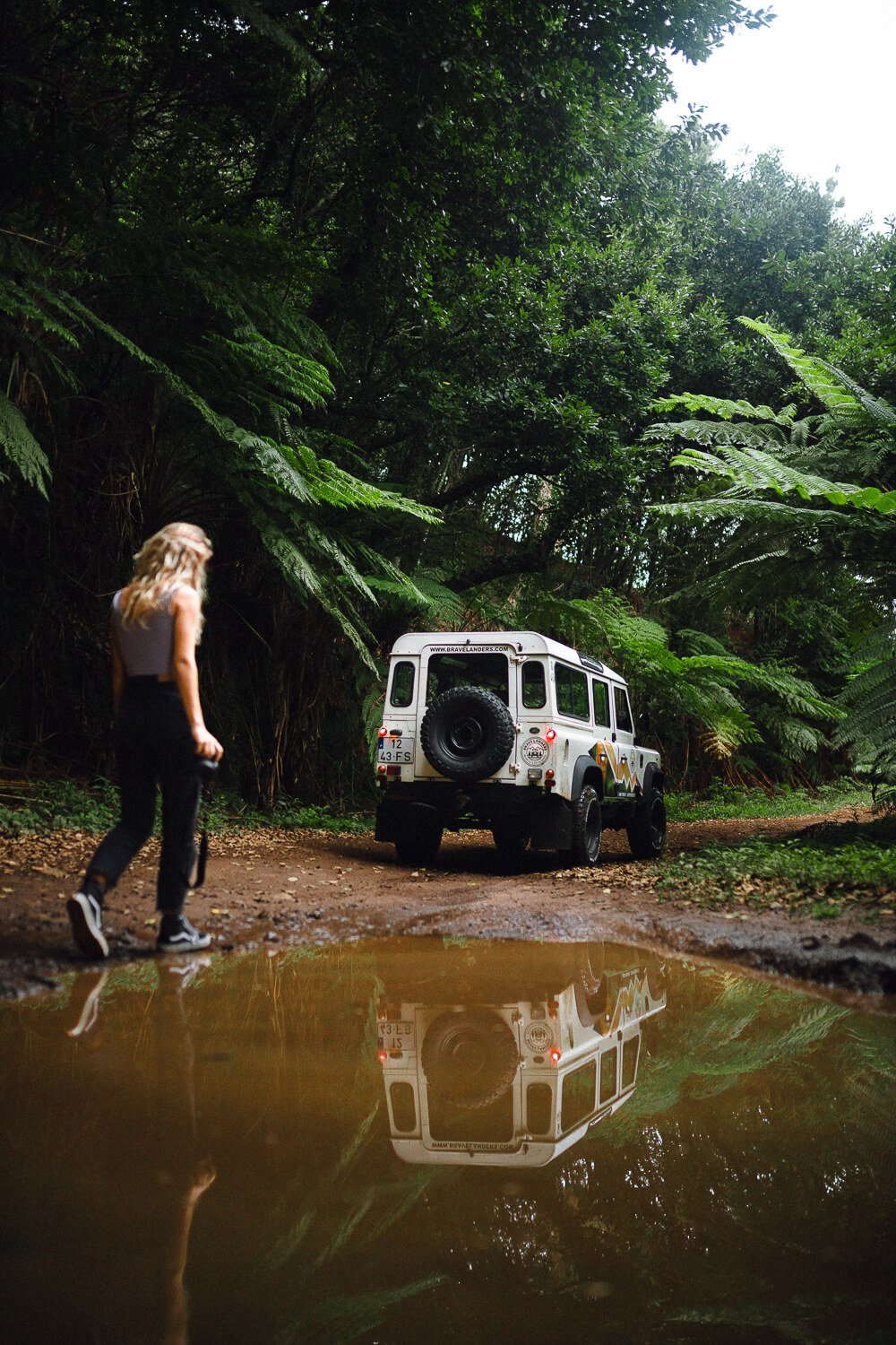 A woman with long blonde hair, wearing a sleeveless white top and black pants, standing on a muddy dirt trail in a lush, green forest, looking at a white off-road vehicle parked on the trail, with a puddle in the foreground reflecting the scene.