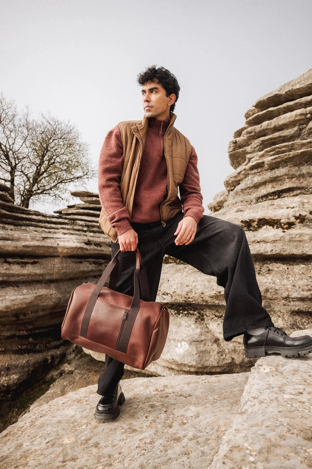 A young man hiking outdoors in a rocky area, dressed in a brown vest over a red sweater, black pants, and black boots, holding a brown duffel bag, looking into the distance with a serious expression.