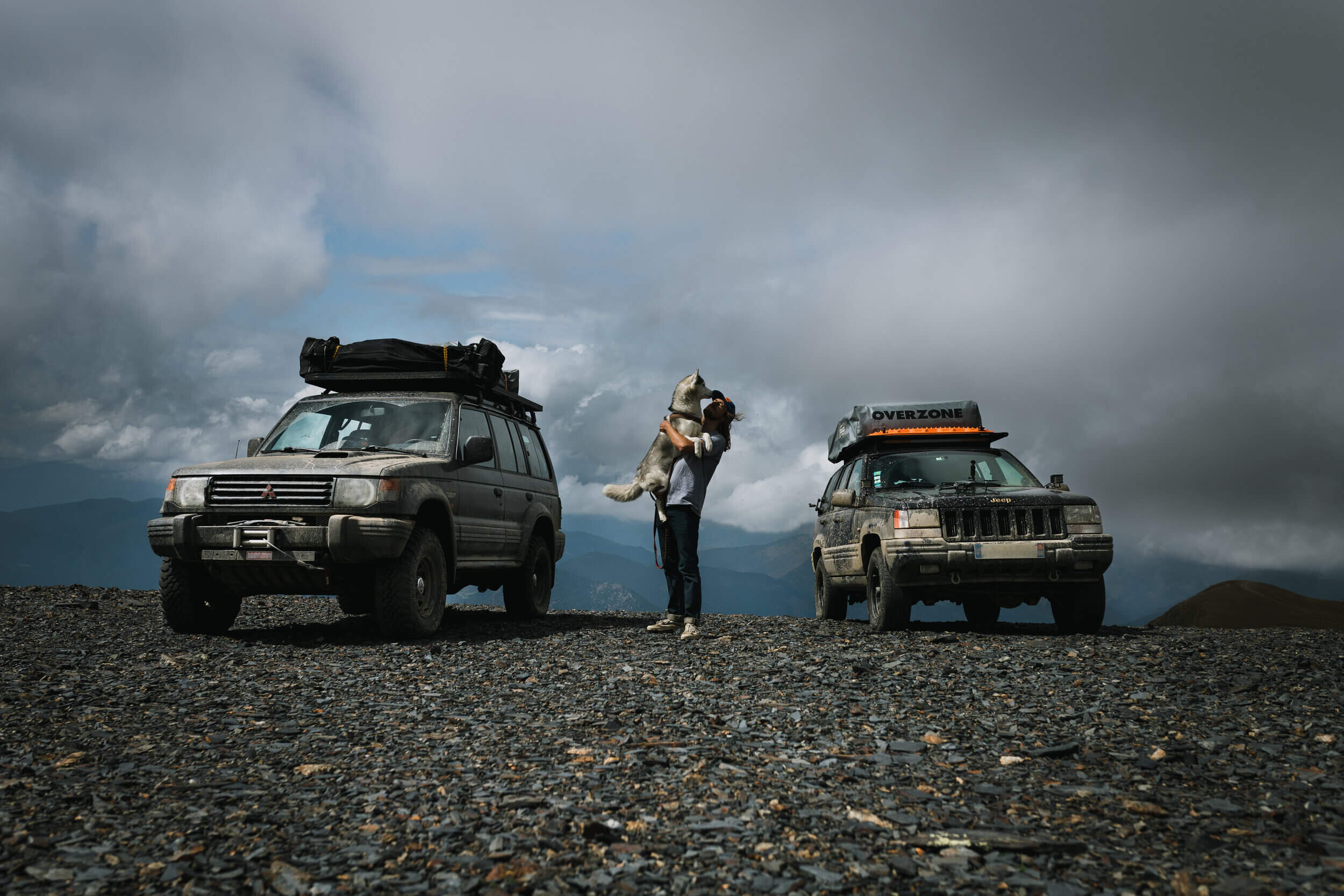 Deux voitures tout-terrain sur un terrain rocheux, avec une personne tenant un chien entre elles. Le ciel est nuageux.