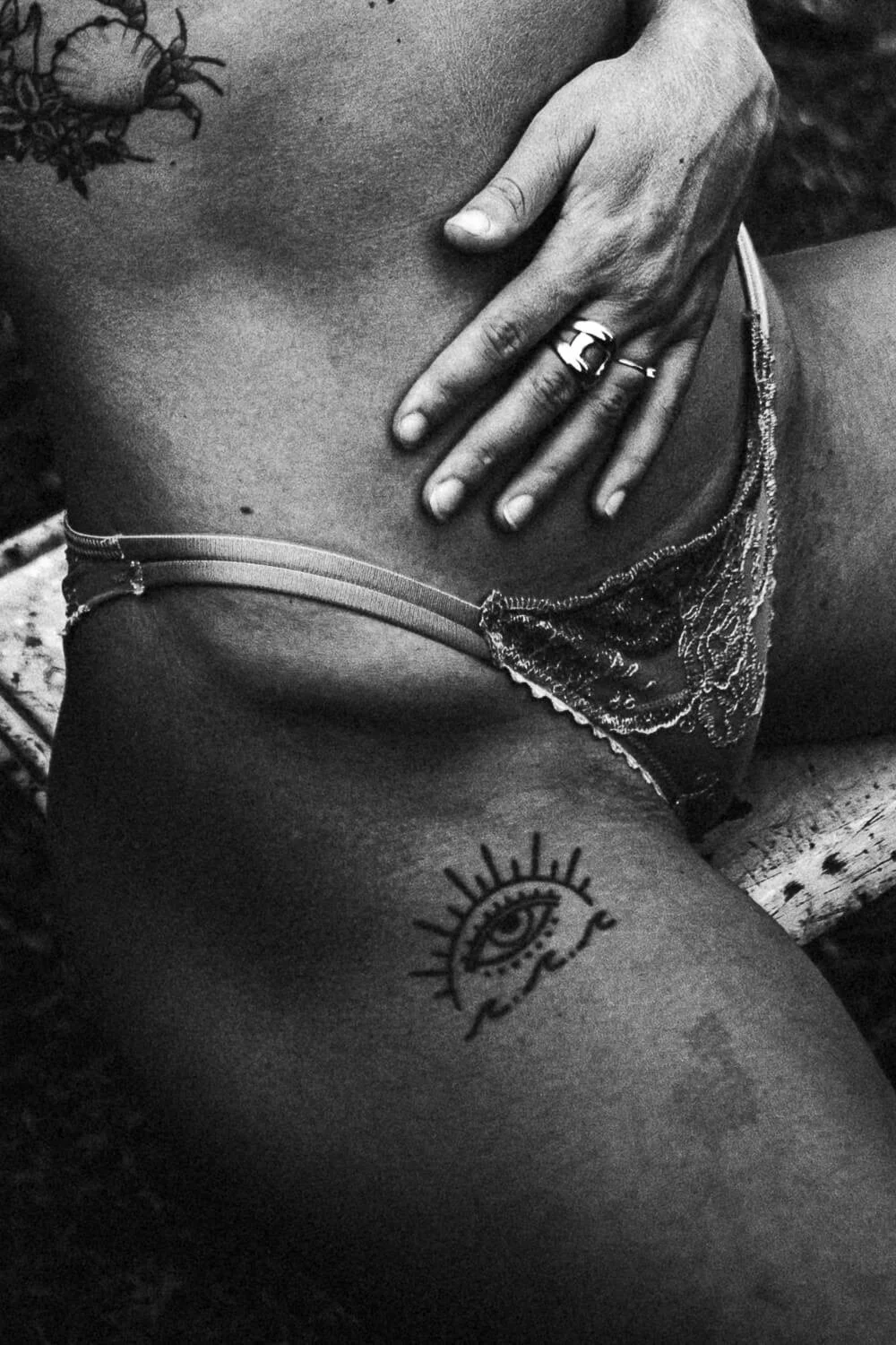 A person’s torso with tattoos, including an eye with rays and a name underneath. The person is wearing lace underwear and a ring, with their hand resting on their lower abdomen.