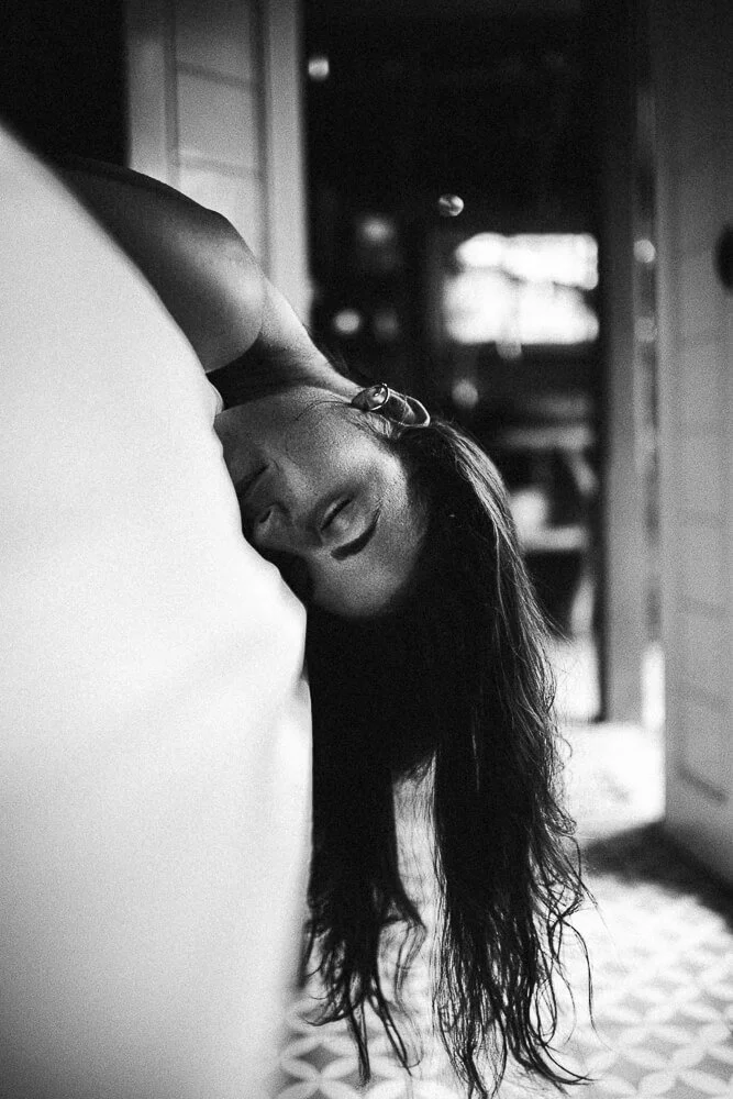 Black and white photo of a woman with long hair leaning on a pillow with her eyes closed, in a room with a window and door in the background.