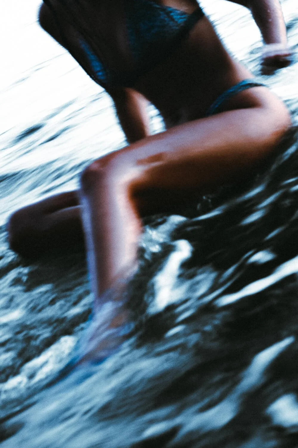 A person in a black top and denim shorts kneeling on water, with their hand touching the water surface. The photo is blurry and taken at an angle.