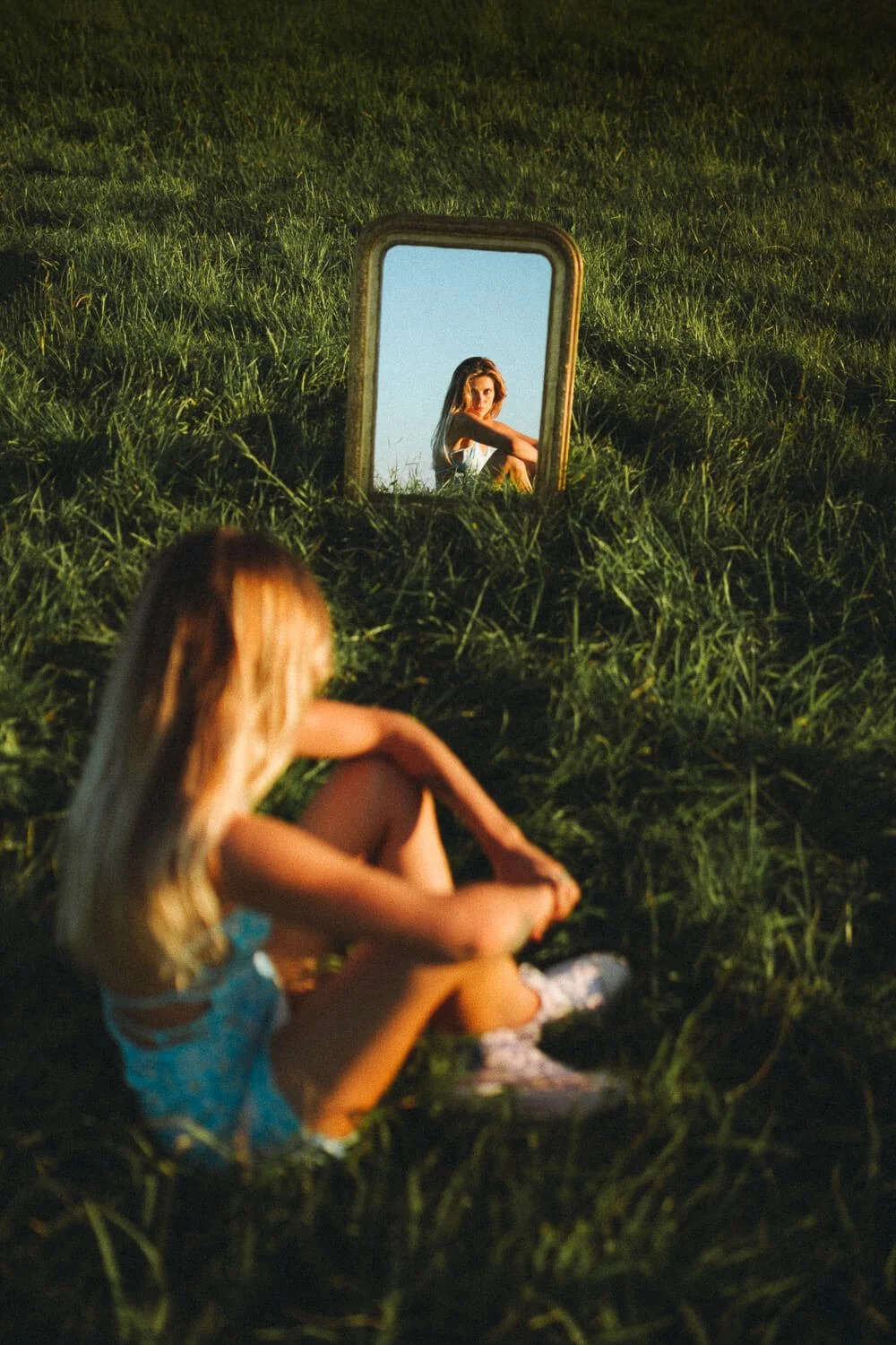 A young girl sitting on green grass, looking into a mirror that shows her reflection.