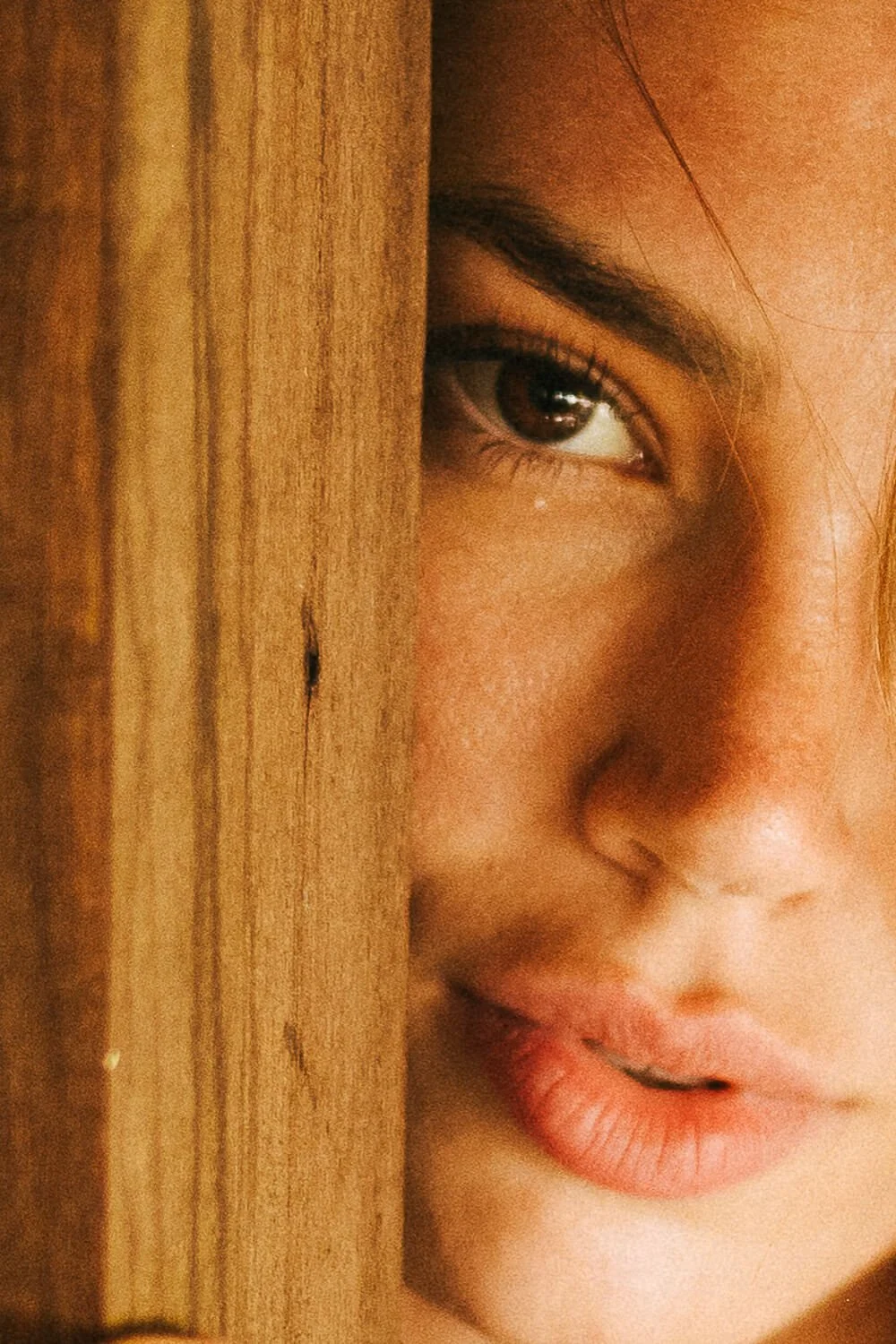A close-up of a child's face peeking out from behind a wooden surface, showing one eye and part of the mouth.