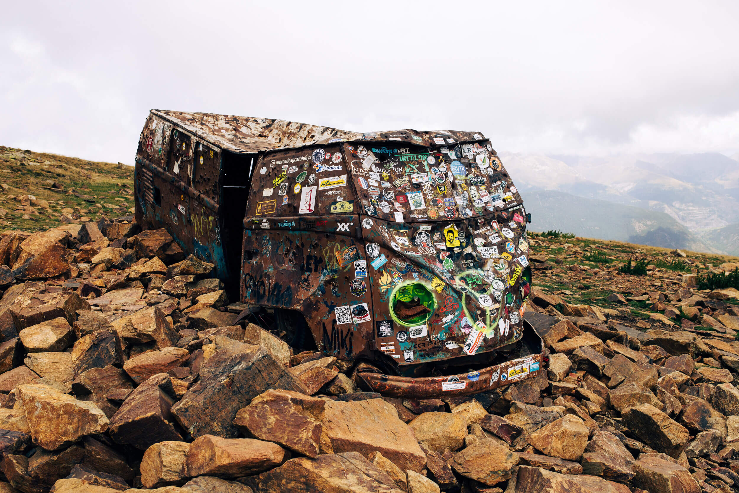 Une vieille carrosserie de Volkswagen Combi rouillée et couverte d'autocollants, posée sur des rochers dans un paysage montagneux.