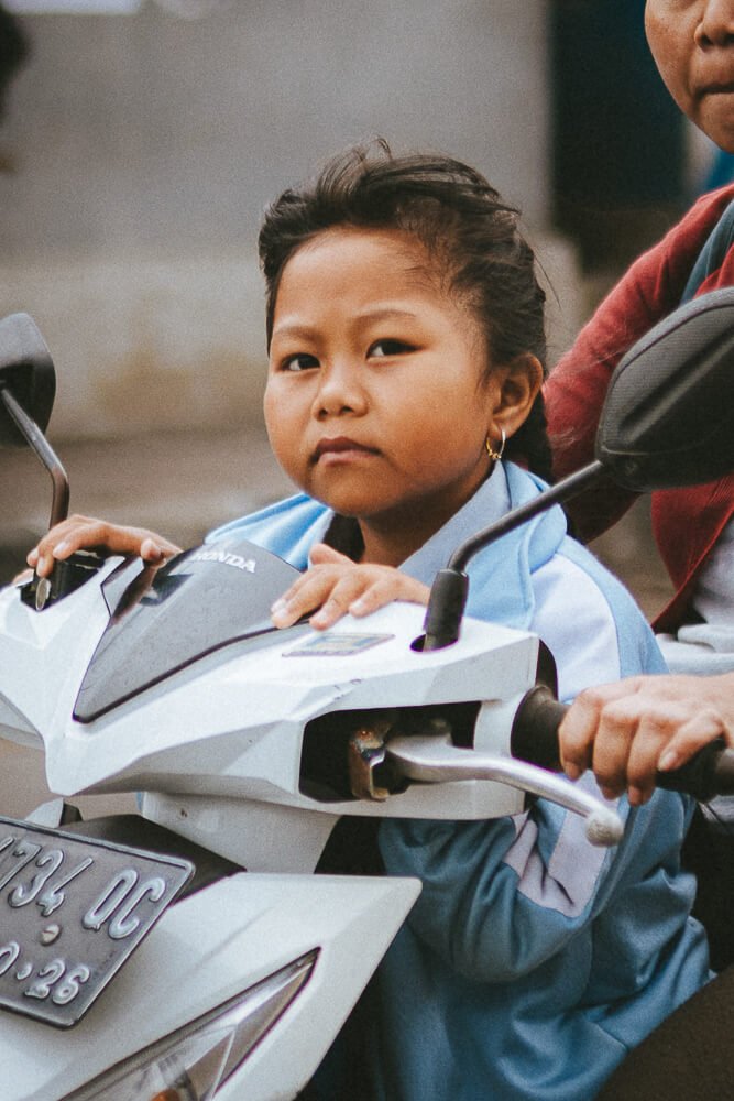 A young girl sitting on a white Honda scooter, holding the handlebars, with a serious expression on her face. She is wearing a light blue jacket and has gold earrings.