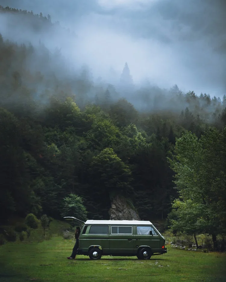 Un homme chargeant le coffre d'une vieille camionnette verte dans un paysage forestier avec des montagnes et de la brume en arrière-plan.