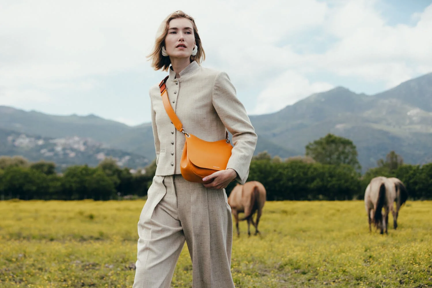 A woman in a light-colored suit stands outdoors in a grassy field with mountains and horses in the background. She carries an orange bag across her shoulder.