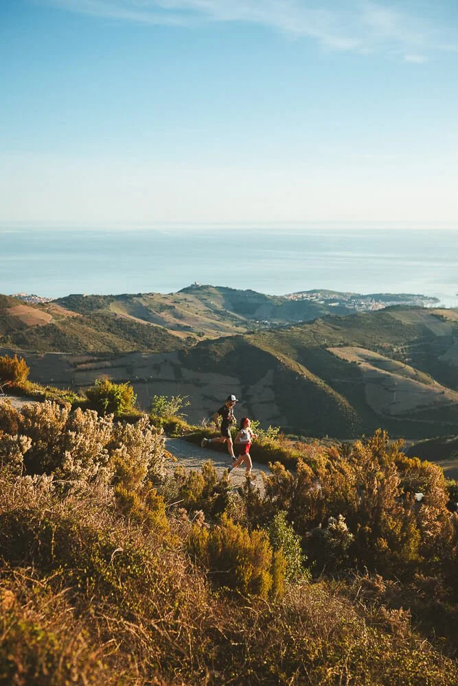 Two people trail running on a mountain trail with lush vegetation, overlooking rolling hills and a distant ocean under a clear sky.